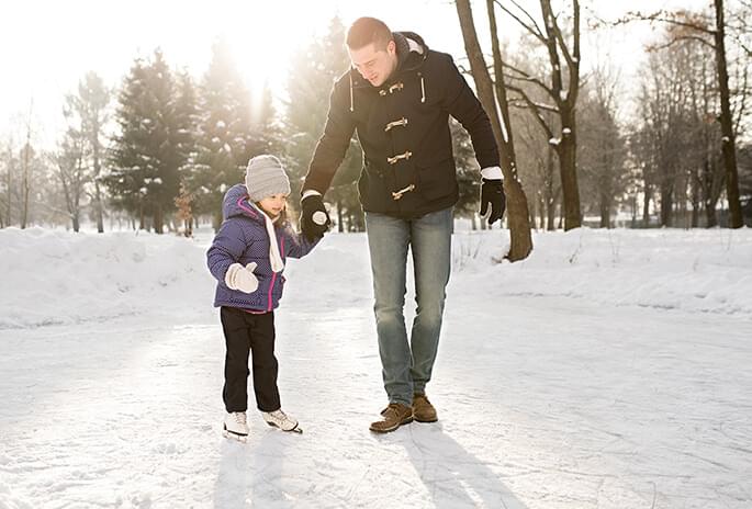 A father holds his daughter's hand as she is learning to ice skate on a pond